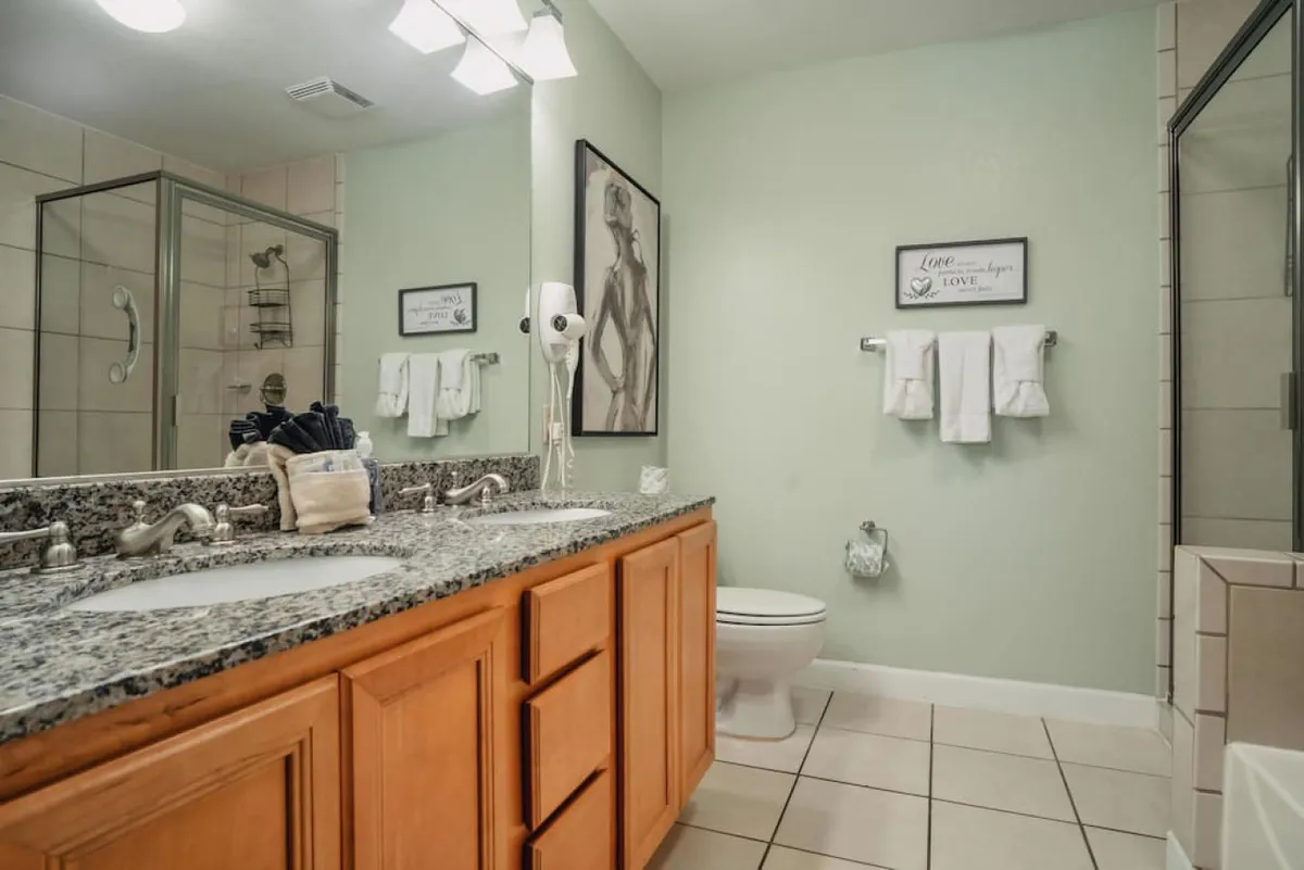A modern bathroom featuring dual sinks with granite countertops, a glass shower enclosure, framed wall art, and neatly hung towels. The space is well-lit and designed for both relaxation and function.
