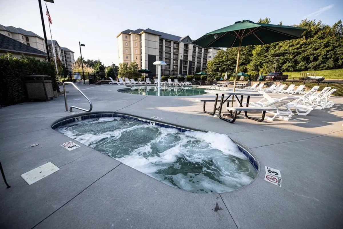 This vibrant exterior image shows a pool area featuring a hot tub, swimming pool, lounge chairs, and umbrellas. The space is designed for relaxation and socializing, with wide walkways suitable for easy movement and accessibility.