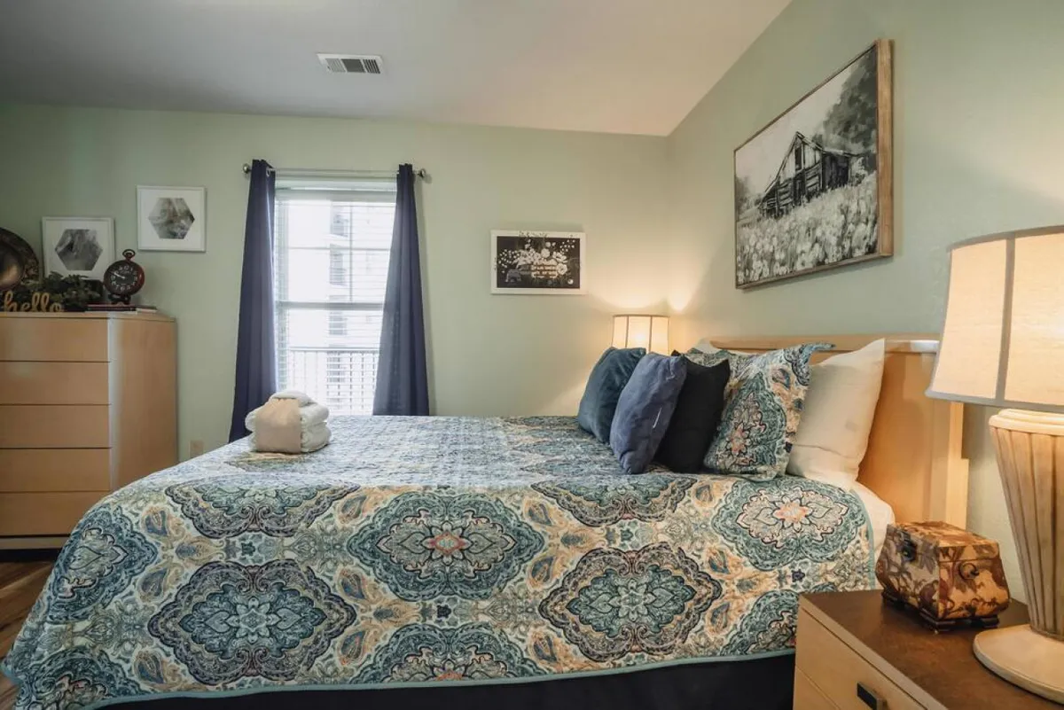 This image shows a cozy bedroom with a bed featuring a patterned quilt, decorative pillows, bedside tables with lamps, and wall art. The room is bathed in natural light, creating a serene and restful ambiance ideal for relaxation.