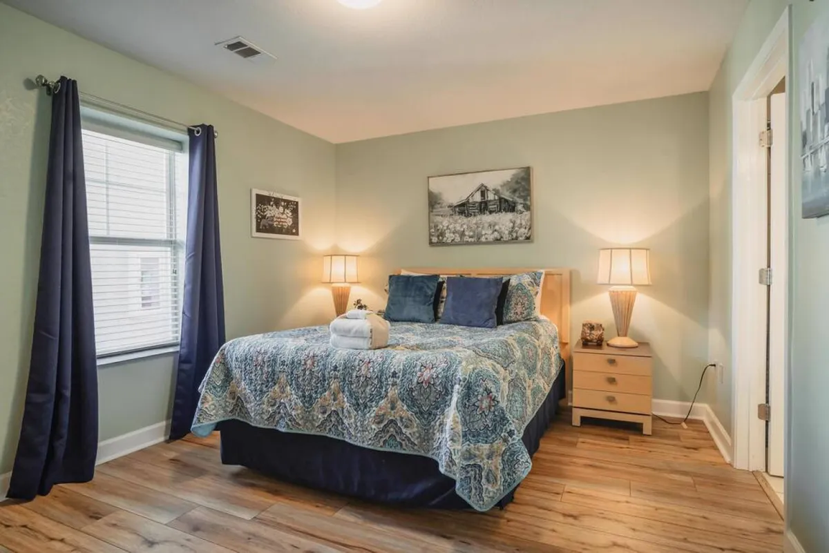 The image features a beautifully appointed bedroom with an inviting bed adorned in ornate bedding, flanked by warm-lit nightstands and soft lamps. Sunlight filters gently through curtained windows, while tasteful artwork completes the serene, welcoming ambiance.