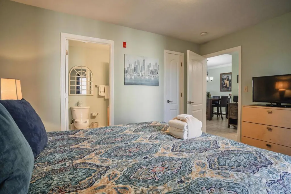 A comfortable bedroom setup with a decorative bed cover, wall art, a television, and a door leading to the bathroom. The space is designed for both relaxation and convenience, making it ideal for unwinding after a busy day.