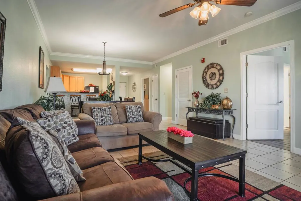 A cozy living room scene with a sofa adorned with decorative pillows, a coffee table with flowers, tasteful wall art, and a view into the adjacent kitchen area. The space is arranged for easy conversation and relaxation, capturing the essence of comfort and connection.
