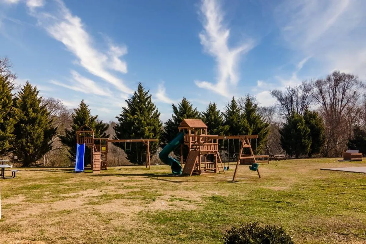 A cheerful playground surrounded by green grass and trees, with children playing and families gathering for picnics, showcasing open spaces and recreational equipment.