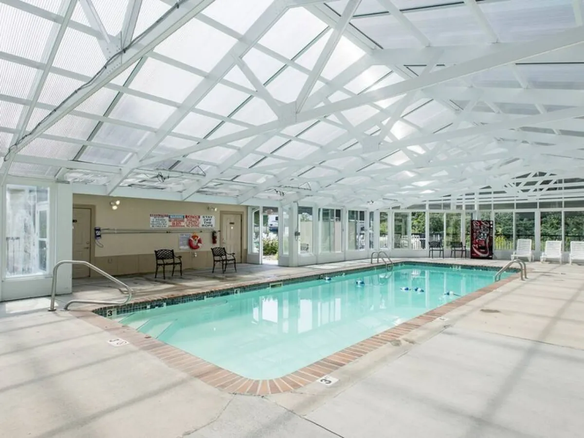 An inviting heated indoor swimming pool with white ceiling beams and lounge chairs lining the side, creating a welcoming atmosphere for swimming and relaxing regardless of the weather.